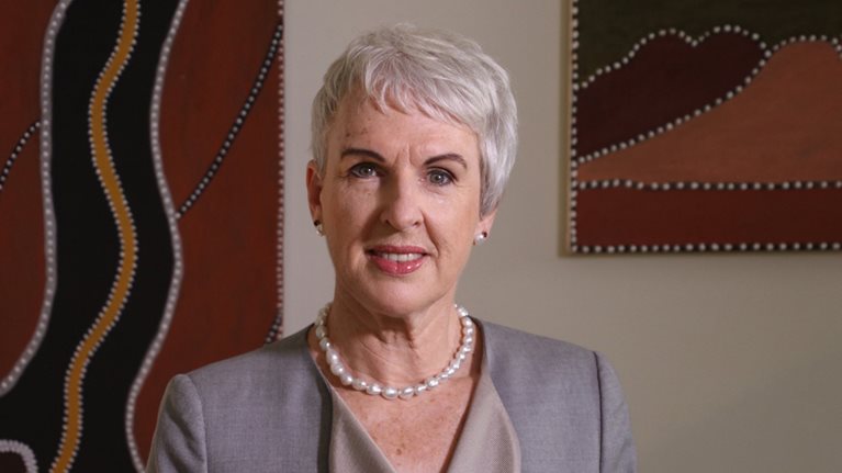 Diane Smith-Gander wearing a light grey suit and a pearl necklace stands in front of abstract artwork on a wall