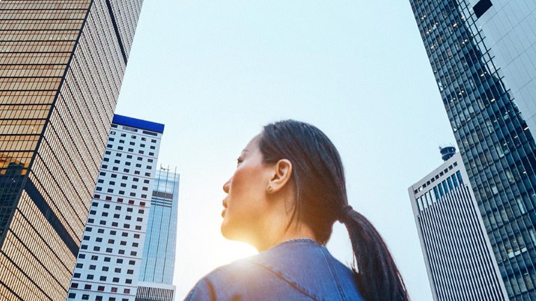 A woman stands with her back to the camera, gazing upward at a cluster of towering skyscrapers that dominate the frame.