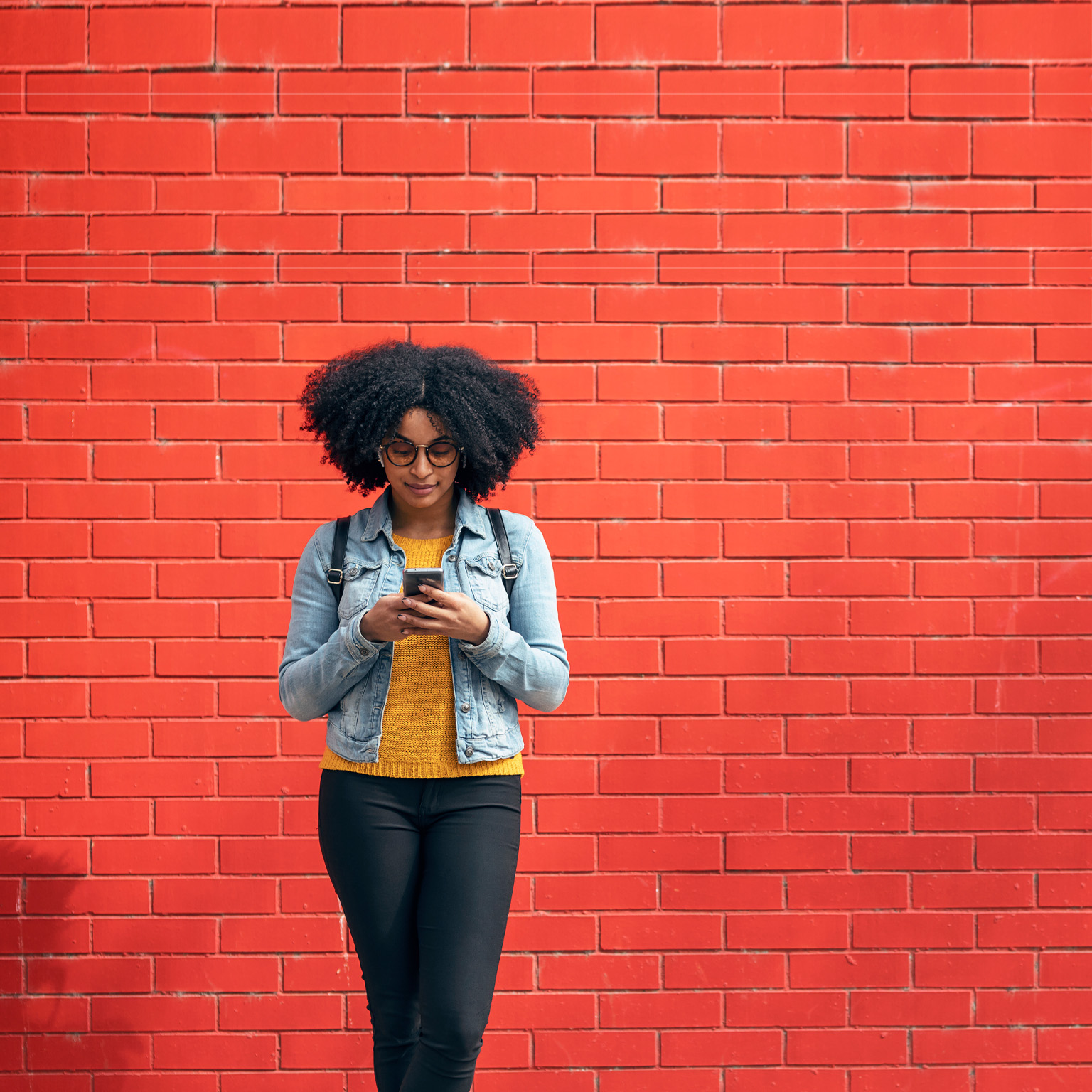 Young woman using smart phone while standing in front of red brick wall - stock photo