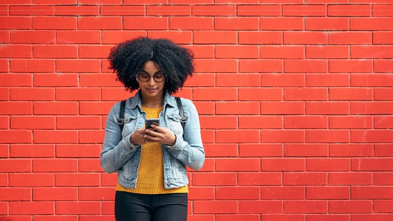 Young woman using smart phone while standing in front of red brick wall - stock photo