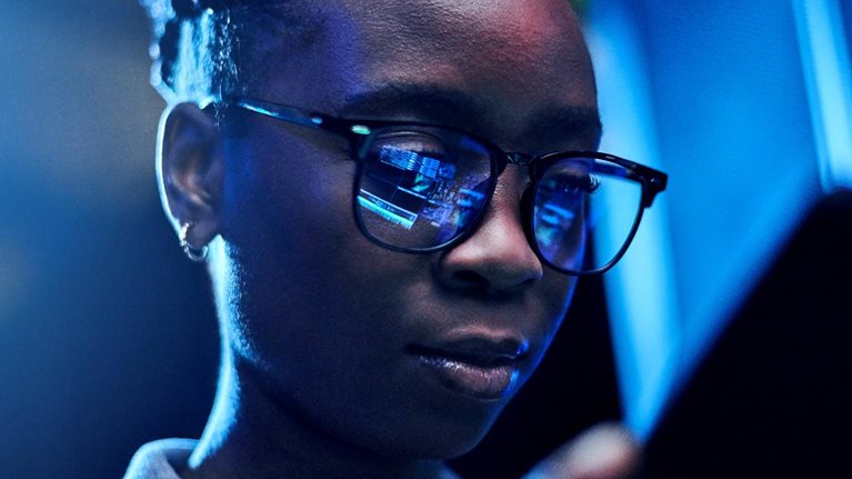 Engineer using a digital tablet while working in a server room with code reflected in her glasses.