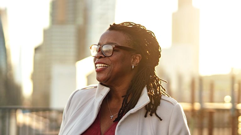A mature businesswoman standing with her hands in pockets on office building rooftop with the London cityscape in the background.