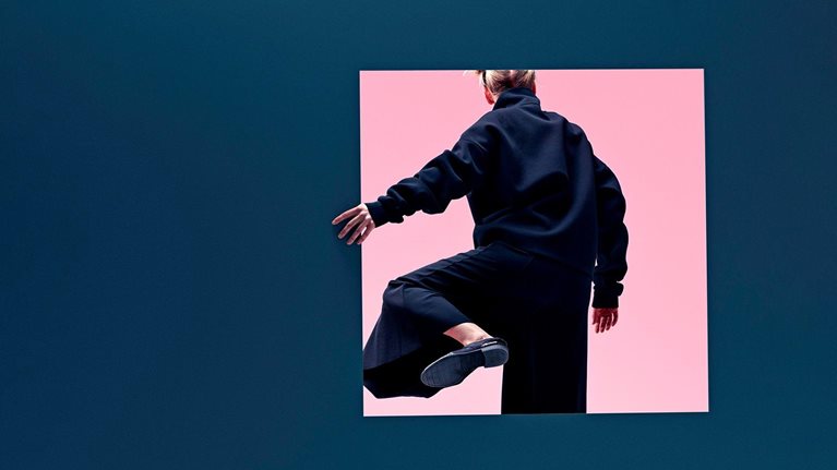 A woman in loose fashionable clothing stepping though a square opening half her height in a dark blue colored wall. The other side of the opening is bright and pink.