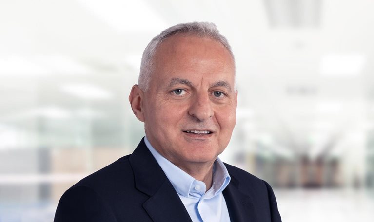 Tufan Erginbilgiç is depicted in a navy blue suit jacket and light blue button-down, smiling against a bright, blurred office setting.