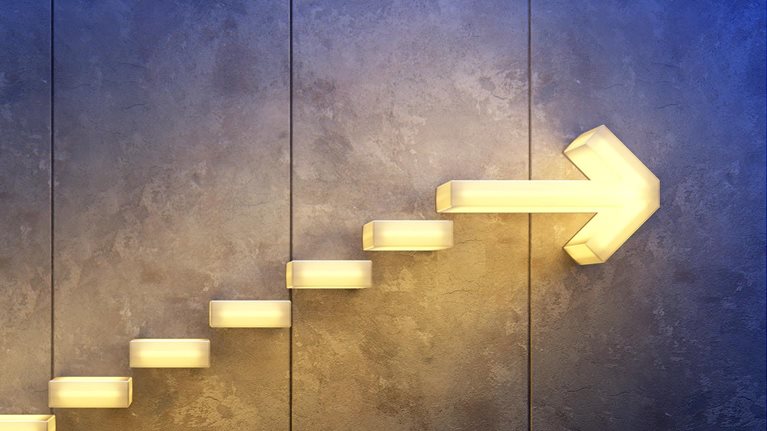 A series of glowing, rectangular stairs arranged in the form of an arrow on a rough concrete surface. The arrow directs upward, indicating progress and accomplishment.