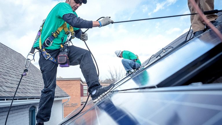 Construction worker uses safety roles to move around a roof with solar panels in the foreground