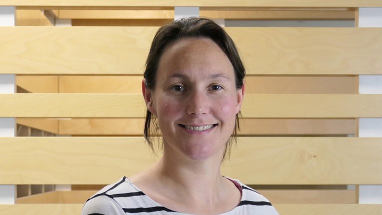 Elspeth Kirkman wearing a white shirt with black stripes, smiling against a backdrop of wood paneling.