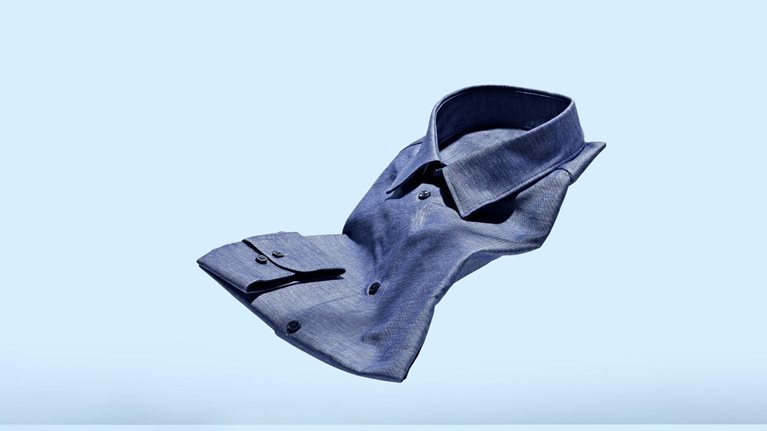 A chambray mens dress shirt floating weightlessly against a light blue background.
