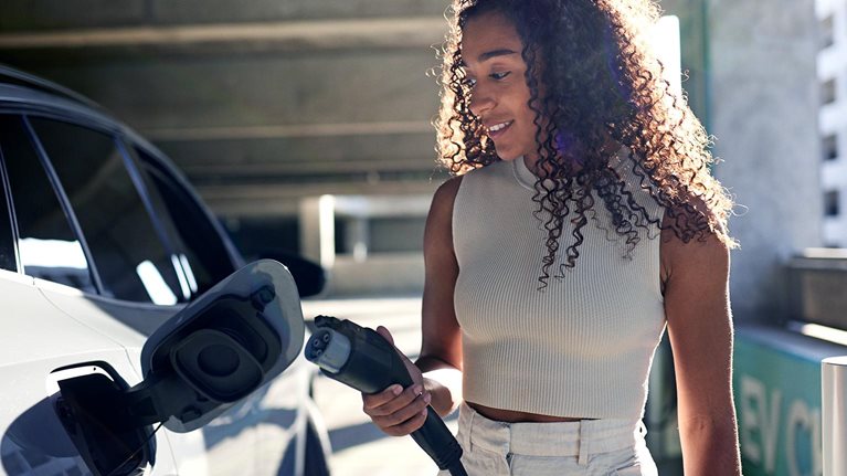 Young woman holding electric plug by car at charging station