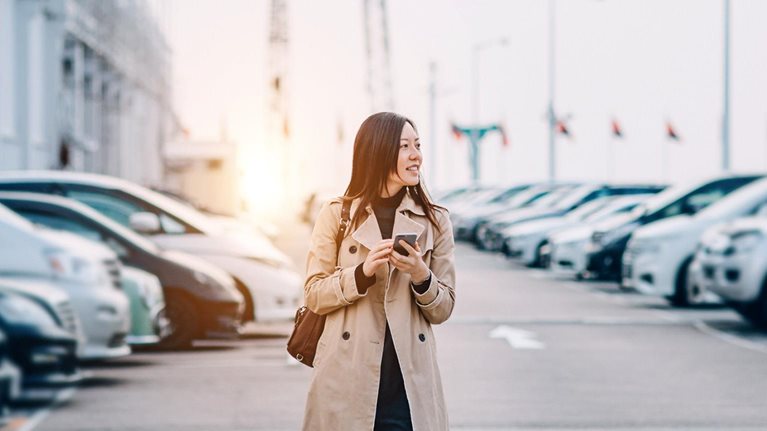 Young lady using smartphone while walking to car in rooftop carpark in city