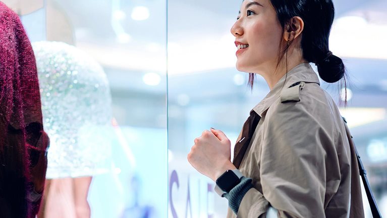 Asian woman carrying a shopping bag, standing outside a boutique looking at window display in a shopping mall