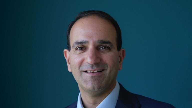 Anant Bhalla wearing a dark suit and a blue dress shirt. His face bears a light smile as he poses against a blue backdrop.