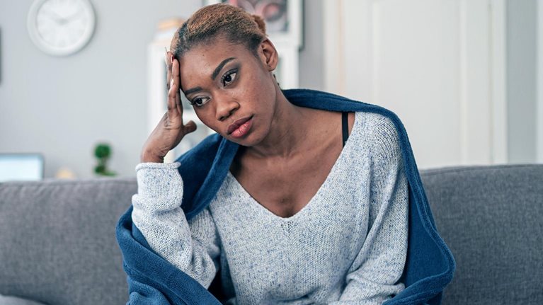 A young woman sitting on her couch wrapped in a blanket with used tissues laying next to her.