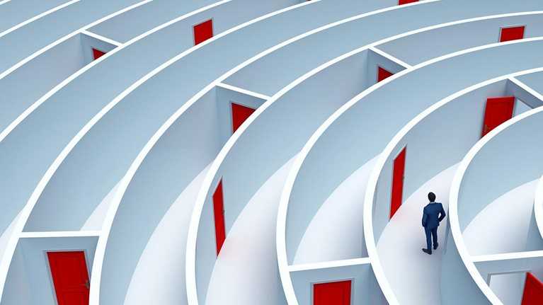 A man stands in the middle of a large maze that has many doors. A pathway out is not clear.