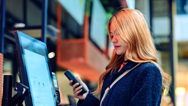 Women in casual clothing holding up her cell phone as she uses a smart checkout screen in a department store.