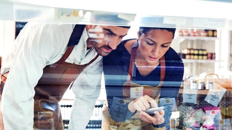 Male and female business owners wearing smocks using smart phone while examining display cabinet in a grocery store