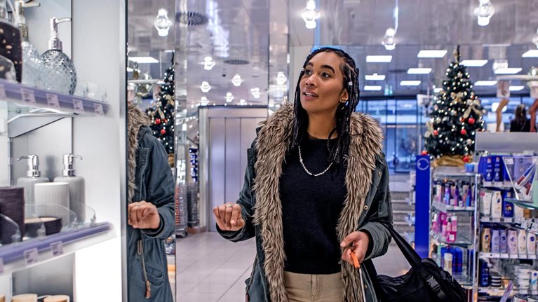 A woman walking down an aisle in a store while looking at personal care items. She is wearing a warm coat and holding a shopping basket, store holiday decorations can be seen in the background.