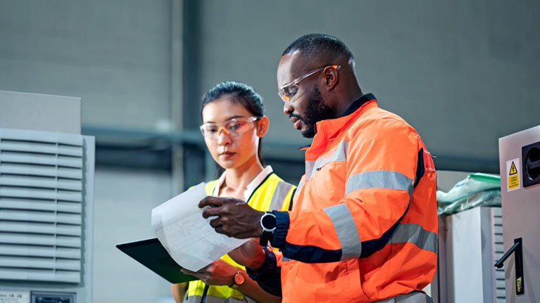 A woman holding a tab and a man with a checklist, both wearing different color-coded work jackets standing in a control room