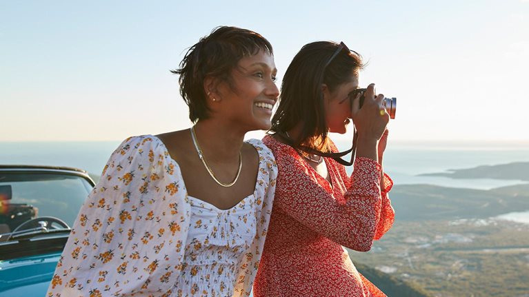 Close up image of friends on vacation sitting on the hood of a car smiling and taking pictures of the view in Montenegro.