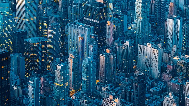 Tight aerial view of skyscrapers in the evening. No horizon line is visible, so the geometric shapes of the buildings and glowing windows of the light-filled offices and apartments become the focus.