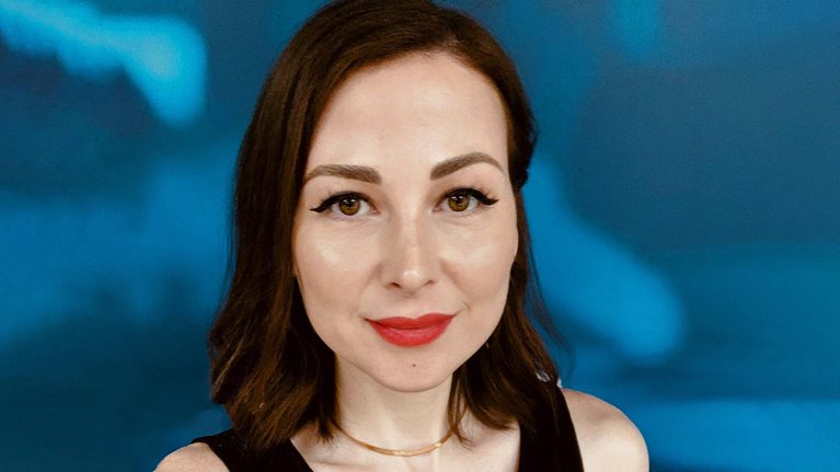 Portrait of Zeinab Hijazi with short dark-brown hair and red lipstick. She is wearing a black tank top and a gold necklace. The background is a blue and white abstract pattern.