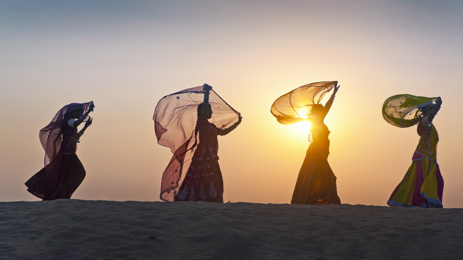 As the sun rises behind them, four Indian women dressed in traditional attire stroll along the crest of a desert dune, casting their figures in silhouette.